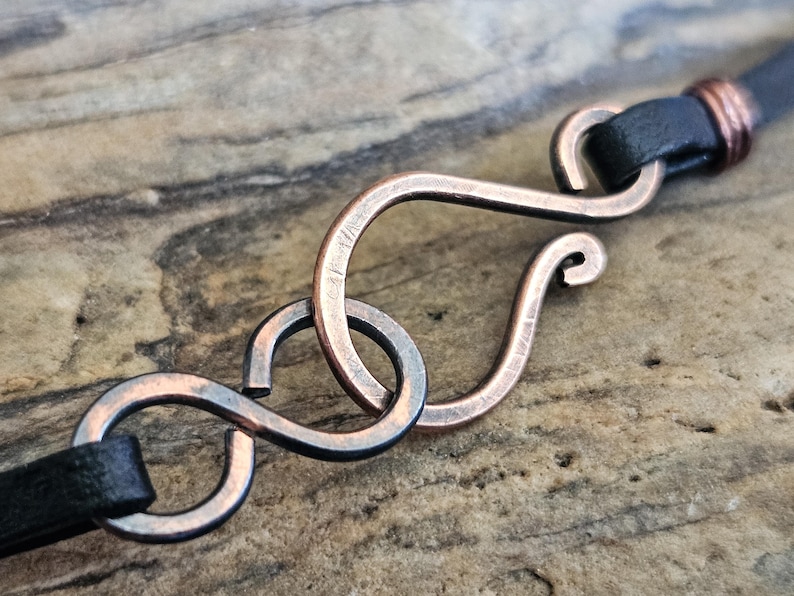 May include: A close-up of a copper-coloured metal clasp with an infinity symbol design. The clasp is attached to a black leather strap.