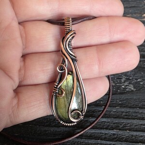 May include: A handcrafted pendant featuring a teardrop-shaped, green and gold stone encased in copper wire. The wire is intricately woven, creating a decorative frame. The pendant hangs from a brown leather cord, showcasing a unique, artisan-made jewelry piece.