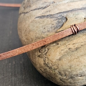 May include: A close-up of a brown leather cord with a copper wire wrap. The cord is lying on a rough, textured rock.