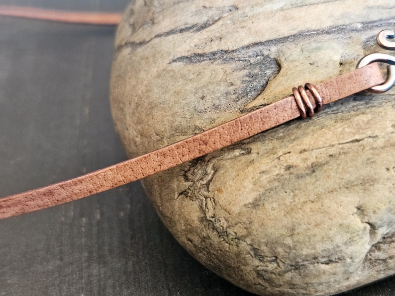 May include: A close-up of a brown leather cord with a copper wire wrap. The cord is lying on a rough, textured rock.