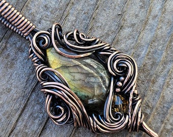 May include: A copper wire wrapped pendant featuring a green and gold labradorite gemstone. The pendant has a delicate, intricate design with swirls and loops.