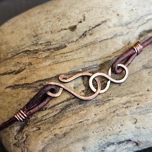 May include: A close-up of a brown leather cord with a copper wire wrap. The cord is attached to a copper wire clasp that is shaped like an infinity symbol.