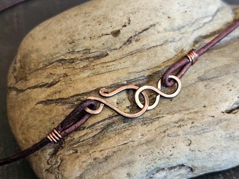 May include: A close-up of a brown leather cord with a copper wire wrap. The cord is attached to a copper wire clasp that is shaped like an infinity symbol.