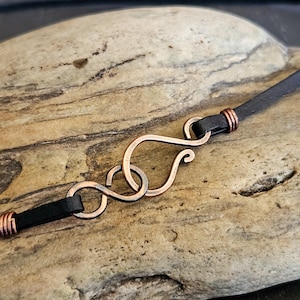 May include: A black leather cord necklace with a copper infinity symbol clasp.