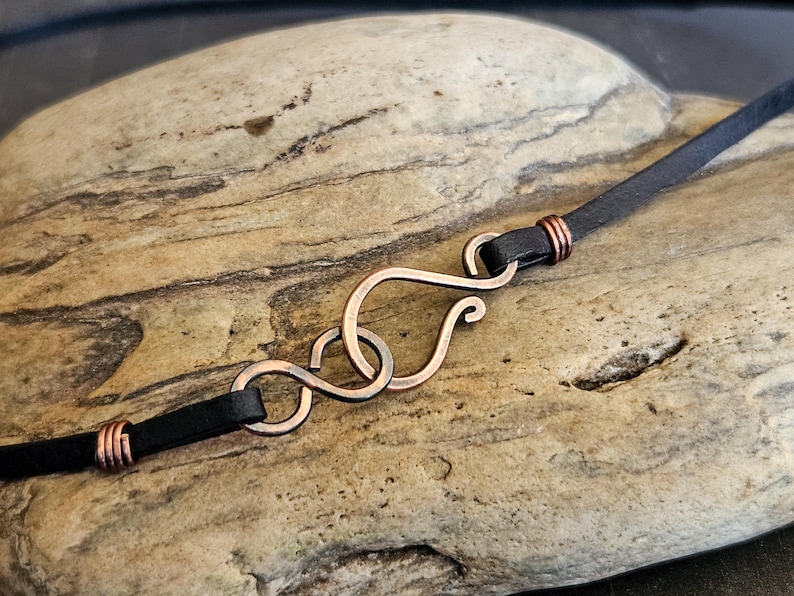 May include: A black leather cord necklace with a copper infinity symbol clasp.