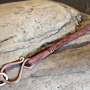 May include: A close-up of a copper clasp attached to a brown leather strap. The clasp is a decorative, ornate design with a loop for attaching to a necklace or bracelet.