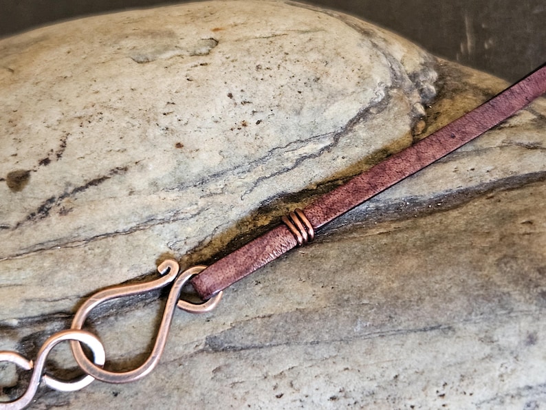 May include: A close-up of a copper clasp attached to a brown leather strap. The clasp is a decorative, ornate design with a loop for attaching to a necklace or bracelet.