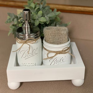 May include: White painted mason jars with a soap dispenser and a sponge on a white tray with wooden legs. The jars are labeled "Ball Mason" and "Ball".