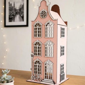 Op de afbeelding: Een hoog, lichtroze houten modelhuis met een trapgevel, een rond zolderraam en meerdere boogramen. De gevel is voorzien van een witte voordeur, een kleine uitgesneden fiets en een lantaarnpaal aan de linkerzijde.