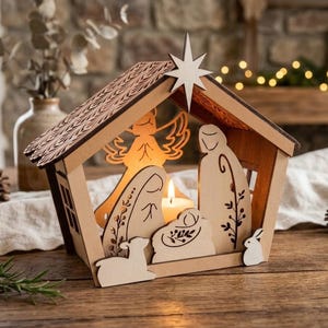 May include: A wooden nativity scene candle holder featuring a laser-cut stable structure with a star on top. Inside, flat wooden silhouettes of figures, an angel, and small animals surround a central lit candle, creating a warm glow.