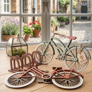May include: Two wooden bicycle models, one light green with a pink seat and one terracotta orange, featuring thin spokes and white-rimmed wheels. A small brown tag tied to the green bicycle with twine displays a tree icon and the text HANDMADE.