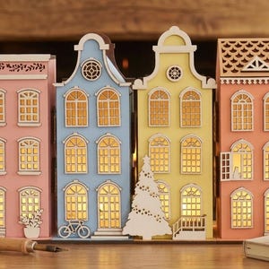 May include: A set of four decorative wooden house structures in pink, blue, yellow, and orange, each featuring intricate cut-out windows with internal warm lighting. The buildings display architectural details like gables, a bicycle, and a tree.