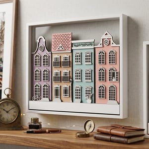 Op de afbeelding: Een witte rechthoekige schaduwdooslijst met een gelaagd houten wandkunstwerk bestaande uit vier kleurrijke miniatuur grachtenpanden in tinten paars, beige, blauwgroen en roze, elk voorzien van gedetailleerde ramen, deuren en daklijnen.