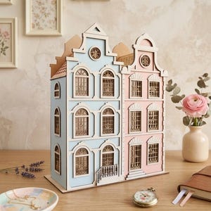 Può includere: Un modello in legno di due case a schiera collegate, una azzurro chiaro e una rosa pallido, con telai delle finestre, porte ad arco e timpani decorativi tagliati al laser. La struttura presenta più livelli con facciate e balconi dettagliati.