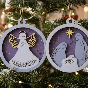 May include: Two circular wooden Christmas ornaments featuring layered cutouts. One ornament displays a white angel with a gold halo and wings against a purple starry background. The other shows a nativity scene with a yellow star above.