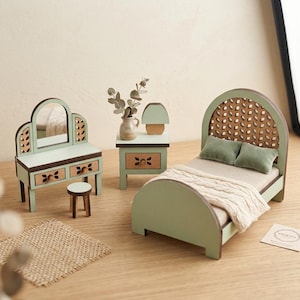 Puede incluir: Un conjunto de muebles de dormitorio en miniatura en tonos verde salvia y madera, que incluye una cama con una manta de punto crema y dos almohadas verdes, un tocador con espejo y taburete, una mesita de noche con lámpara y un jarrón con follaje.