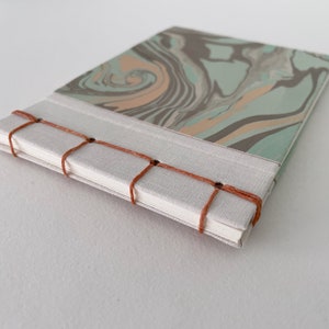 May include: A handmade journal with a marbled cover in shades of gray, teal, and peach. The spine is bound with a light beige fabric and stitched with orange thread. The journal is closed.