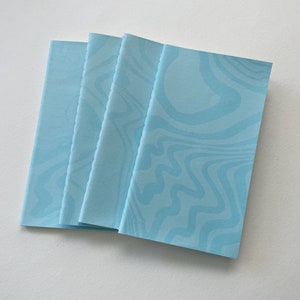 May include: Four light blue notebooks with a wavy pattern in a slightly darker shade of blue. The notebooks are stacked on top of each other, with the top one slightly fanned out. The background is a plain white surface.