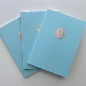 May include: Three light blue notebooks are arranged on a white surface. Each notebook has a circular sticker with a colorful, marbled design in shades of pink, yellow, and gray. The notebooks are slightly angled, showcasing their covers.
