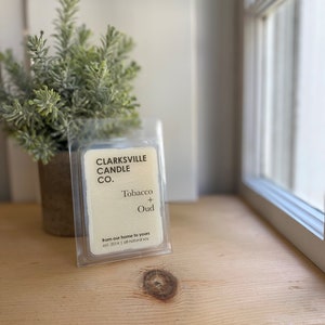 May include: A clear plastic container with a white label that reads "CLARKSVILLE CANDLE CO." and "Tobacco + Oud". The label also includes the text "from our home to yours est. 2014 all-natural soy".