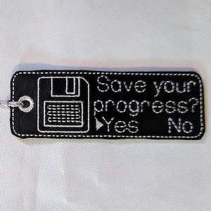 Può includere: Segnalibro nero con ricamo bianco di un floppy disk e il testo "Save your progress? Yes No". Il segnalibro ha un fiocco bianco.