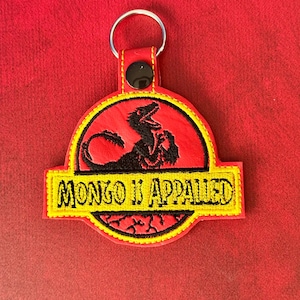 Può includere: Un portachiavi ricamato circolare con sfondo rosso e una sagoma nera di dinosauro. Una fascia orizzontale gialla al centro mostra il testo nero "MONGO IS APPALLED". Il design include un anello di metallo e un bottone a pressione nero.