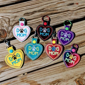 May include: Seven heart-shaped keychains with the text "Dog Mom" and a paw print. The keychains are in various colors, including yellow, teal, red, purple, black, and white.
