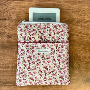 May include: A quilted fabric sleeve featuring a floral pattern of pink roses and green leaves on a cream background. The sleeve has a zipper closure and a white label with the text SMASHCRAFT. A white e-reader is partially inserted, displaying the text Great on Kindle.