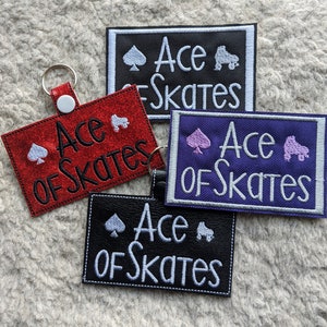 Może przedstawiać: Cztery breloki z napisem "Ace of Skates" w białym kolorze na czarnym, czerwonym, fioletowym i czarnym tle. Każdy brelok zawiera białą lub r&oacute;żową pikę i białe rolki.