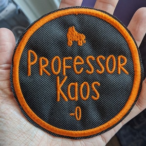Może przedstawiać: Naszywka haftowana w kolorze czarnym i pomarańczowym z napisem "Professor Kaos -0" i małym wzorem rolek.