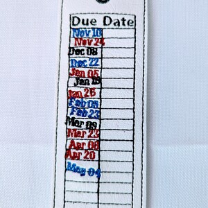 May include: White leather bookmark with a stitched grid and embroidered text that reads "Due Date" and a list of dates in blue and red. The dates are: Nov 10, Nov 24, Dec 08, Dec 22, Jan 05, Jan 15, Jan 26, Feb 09, Feb 23, Mar 08, Mar 23, Apr 08, Apr 20, May 04.