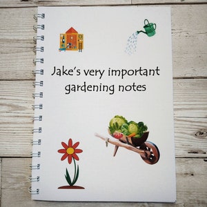 Könnte beinhalten: Ein weißes Spiralnotizbuch mit dem Text "Jake's very important gardening notes" auf dem Einband. Der Einband zeigt außerdem Illustrationen eines Schuppens, einer Gießkanne, einer Schubkarre voller Gemüse und einer roten Blume.