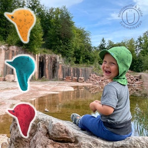 Könnte beinhalten: Ein Kind trägt einen grün-weißen gemusterten Sonnenhut mit dem Text "Merinowolle & Seide Sommerkappe" und sitzt auf einem großen Felsen. Im Hintergrund sind drei verschiedene farbige Sonnenhüte zu sehen.
