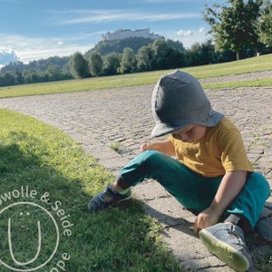 May include: A young child wearing a gray hat and a yellow shirt sits on a cobblestone path. The child is wearing blue pants and has their legs crossed. The child is looking down at something in their hands.