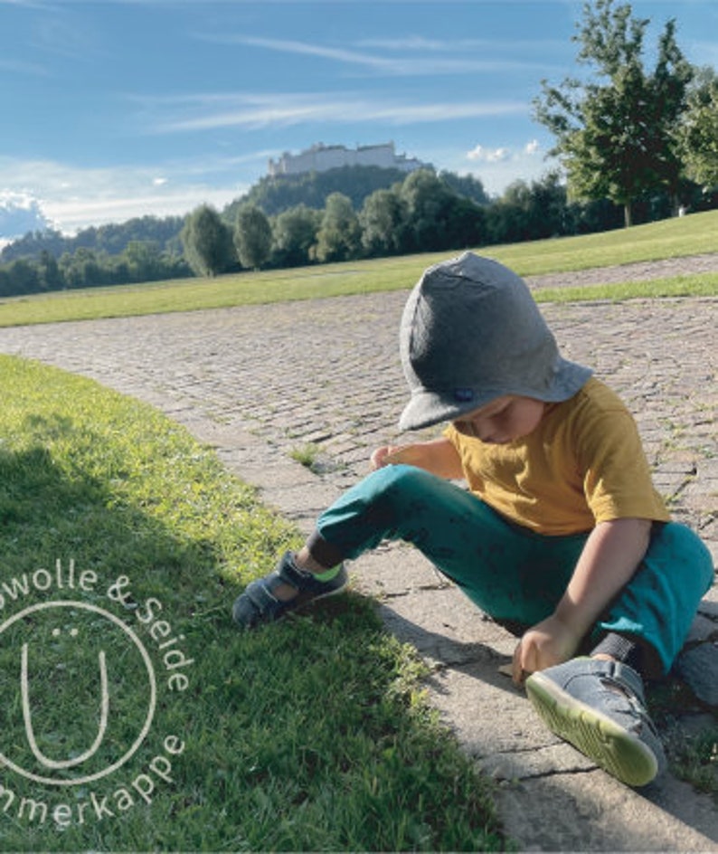 May include: A young child wearing a gray hat and a yellow shirt sits on a cobblestone path. The child is wearing blue pants and has their legs crossed. The child is looking down at something in their hands.