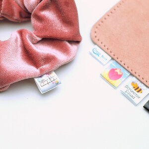 May include: A close-up of a dusty rose velvet scrunchie with a small tag that reads "little fifi handmade." Several other tags are visible, including one that says "Scrunchie Squad" with a cartoon bee.