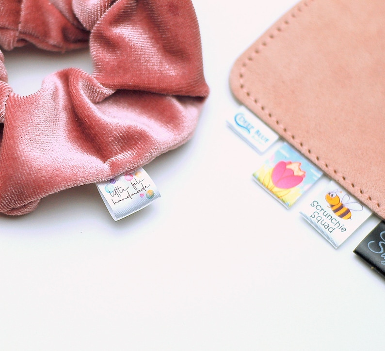 May include: A close-up of a dusty rose velvet scrunchie with a small tag that reads "little fifi handmade." Several other tags are visible, including one that says "Scrunchie Squad" with a cartoon bee.