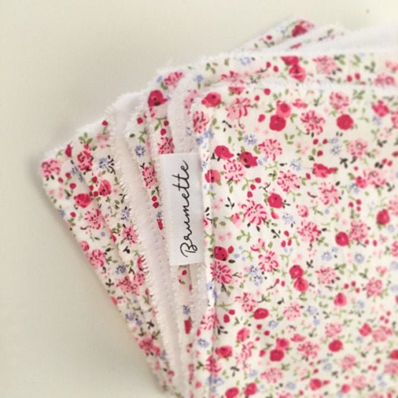 K&ouml;nnte beinhalten: Ein Stapel wei&szlig;er Baumwollstoffe mit einem rosa- und blauen Blumendruck. Die Stoffe haben ein wei&szlig;es Etikett mit dem Wort "Broumette" darauf.