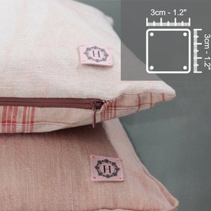 Puede incluir: Dos fundas de cojín de lino rosa y blanco con ribete de vichy rojo y blanco. Cada funda de cojín tiene una pequeña etiqueta de tela con la letra "H" en un círculo.