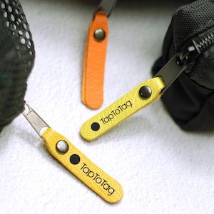Könnte beinhalten: Nahaufnahme von drei Reißverschluss-Anhängern mit Lederlaschen. Zwei sind gelb mit dem Text "TapToTag" und einem schwarzen Punkt. Einer ist orange. Alle haben Metallbeschläge und sind an schwarzen Taschen befestigt.