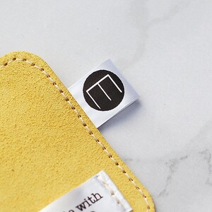 May include: A close-up shot of a yellow suede leather item with a stitched edge. A white tag with a black circular logo featuring the letter "E" is attached. The background is a light, marbled surface.
