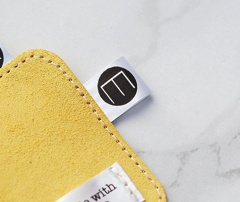May include: A close-up shot of a yellow suede leather item with a stitched edge. A white tag with a black circular logo featuring the letter "E" is attached. The background is a light, marbled surface.