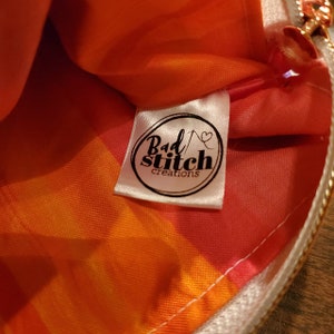 K&ouml;nnte beinhalten: Nahaufnahme einer wei&szlig;en Stofftasche mit Rei&szlig;verschluss. Die Tasche hat ein rot-orangenes Batik-Muster und ein wei&szlig;es Etikett mit dem Text "Bad Stitch creations" in Schwarz.
