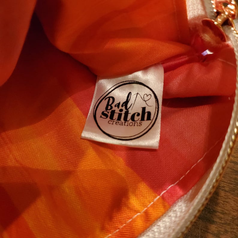 K&ouml;nnte beinhalten: Nahaufnahme einer wei&szlig;en Stofftasche mit Rei&szlig;verschluss. Die Tasche hat ein rot-orangenes Batik-Muster und ein wei&szlig;es Etikett mit dem Text "Bad Stitch creations" in Schwarz.