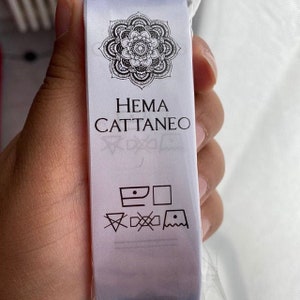 K&ouml;nnte beinhalten: Wei&szlig;es Kleidungsetikett mit schwarzem Text und einem Mandala-Design. Der Text lautet "HEMA CATTANEO" und enth&auml;lt Pflegehinweise.