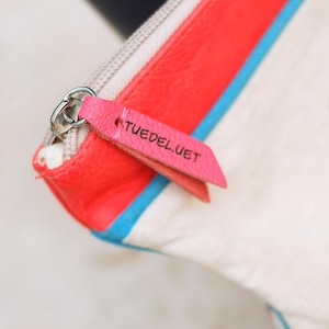 Puede incluir: Primer plano de un bolso blanco con un diseño de rayas rojas y azules. Una etiqueta de cuero rosa con el texto "TUEDEL.UET" está sujeta a la cremallera plateada.