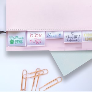 K&ouml;nnte beinhalten: Ein Satz von f&uuml;nf wei&szlig;en Satin-Etiketten mit unterschiedlichen Texten und Designs. Die Etiketten sind horizontal auf einer rosa Oberfl&auml;che angeordnet. Der Text auf den Etiketten lautet "Prettiest Paws", "bibs and hugs", "Nana", "Chouchouette & Patatouille" und "KHOIKHOI LONDON".