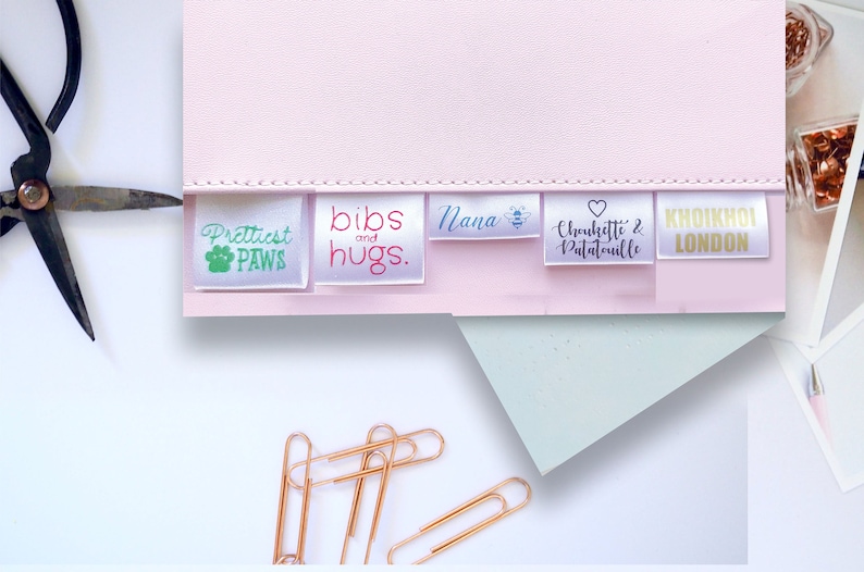 K&ouml;nnte beinhalten: Ein Satz von f&uuml;nf wei&szlig;en Satin-Etiketten mit unterschiedlichen Texten und Designs. Die Etiketten sind horizontal auf einer rosa Oberfl&auml;che angeordnet. Der Text auf den Etiketten lautet "Prettiest Paws", "bibs and hugs", "Nana", "Chouchouette & Patatouille" und "KHOIKHOI LONDON".