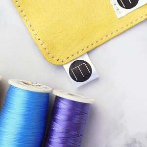 May include: A close-up image shows a yellow suede item with white stitching and a white tag with a black circular logo. Three spools of thread in yellow, blue, and purple are in the foreground, suggesting a crafting theme.