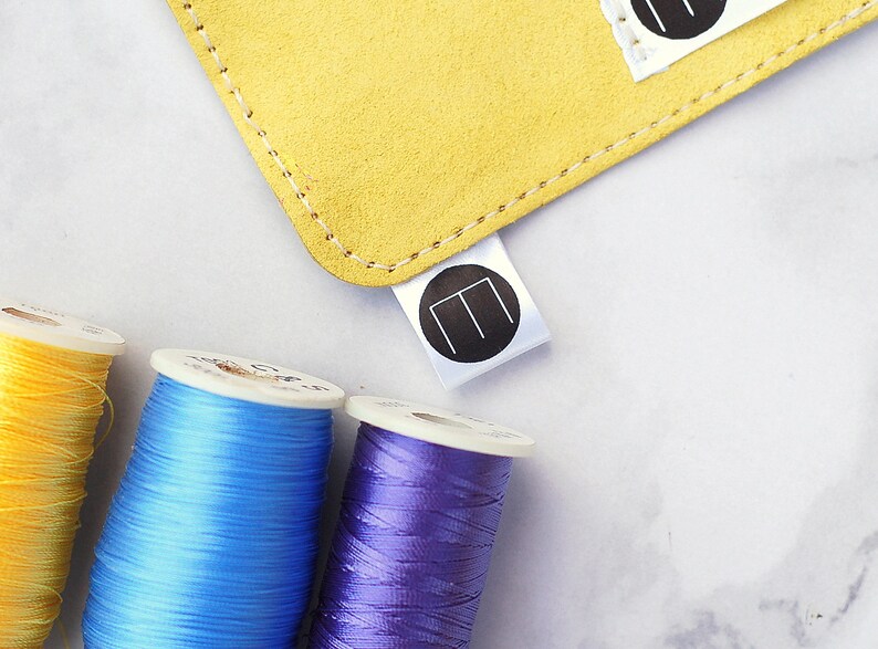 May include: A close-up image shows a yellow suede item with white stitching and a white tag with a black circular logo. Three spools of thread in yellow, blue, and purple are in the foreground, suggesting a crafting theme.